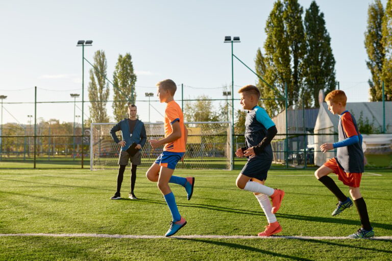 Three boys run a conditioning drill on a turf field while their coach watches with a clipboard.