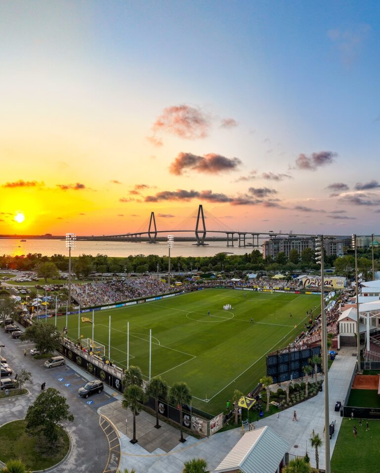 An aerial view of a packed soccer stadium at sunset with the Ravenel Bridge in the background.