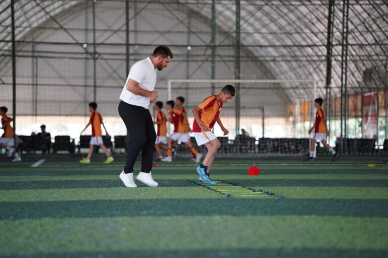 A coach jogs alongside a young player doing footwork through an agility ladder in an indoor facility.