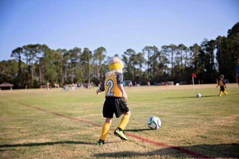A youth player in a yellow jersey sprints after the ball on a sunlit field with teammates in the distance.