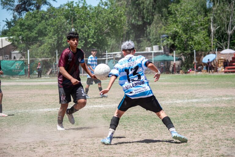 Two teenage soccer players battle for the ball on a dusty field with sponsor banners in the background.