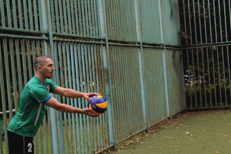 A soccer player in a green jersey leans back to take a throw-in next to a weathered metal fence.