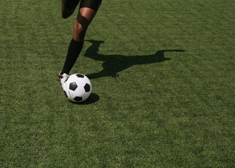 Close-up of a soccer player's leg and foot rolling a ball across turf, casting a long shadow.