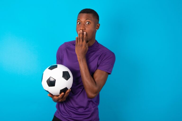 A man in a purple jersey covers his mouth in mock surprise while holding a soccer ball against a blue backdrop.