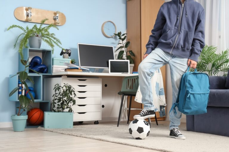 A teen boy balances a soccer ball underfoot while holding a backpack in a living room.