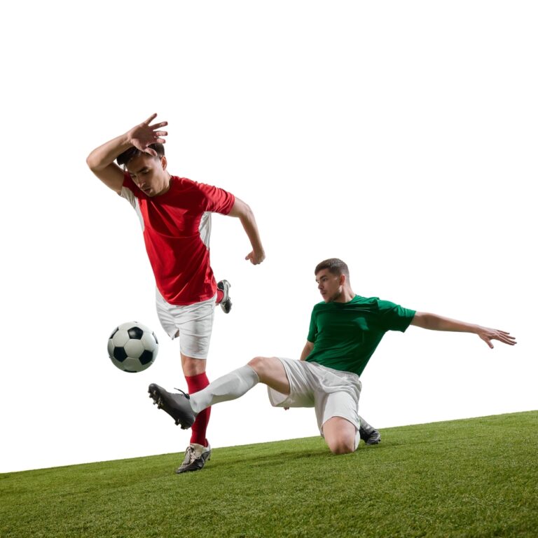 A player in red kicks the ball away as an opponent in green slides in for a tackle on a patch of grass.
