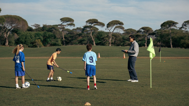 A coach holding a clipboard watches three kids practice dribbling past agility poles on a grass field.