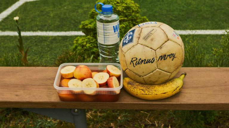 A water bottle, sliced fruit, and a worn soccer ball with a banana sit on a wooden bench by a field.