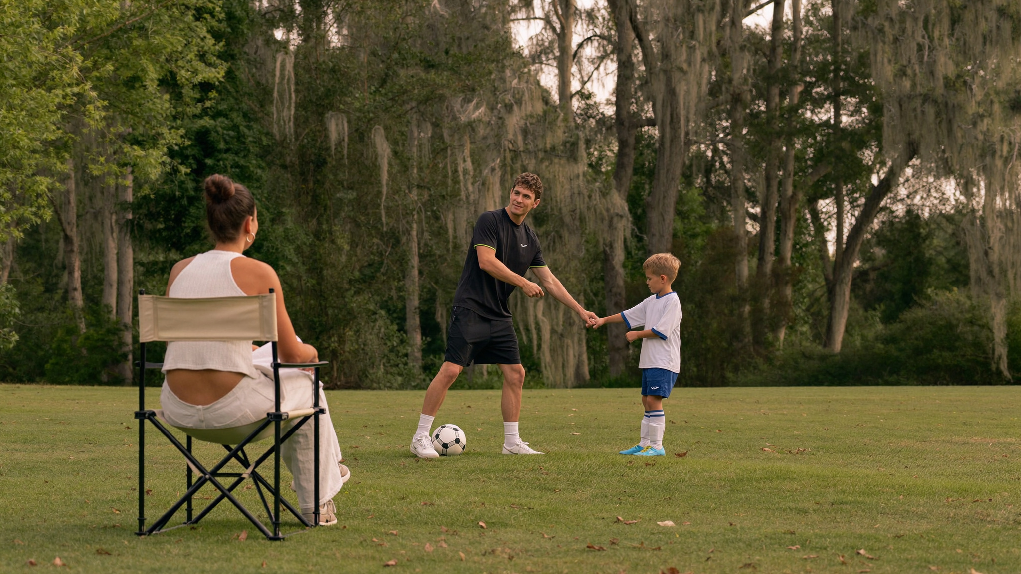 A mother watches from a sideline chair as a coach high-fives a young boy on a grassy field.