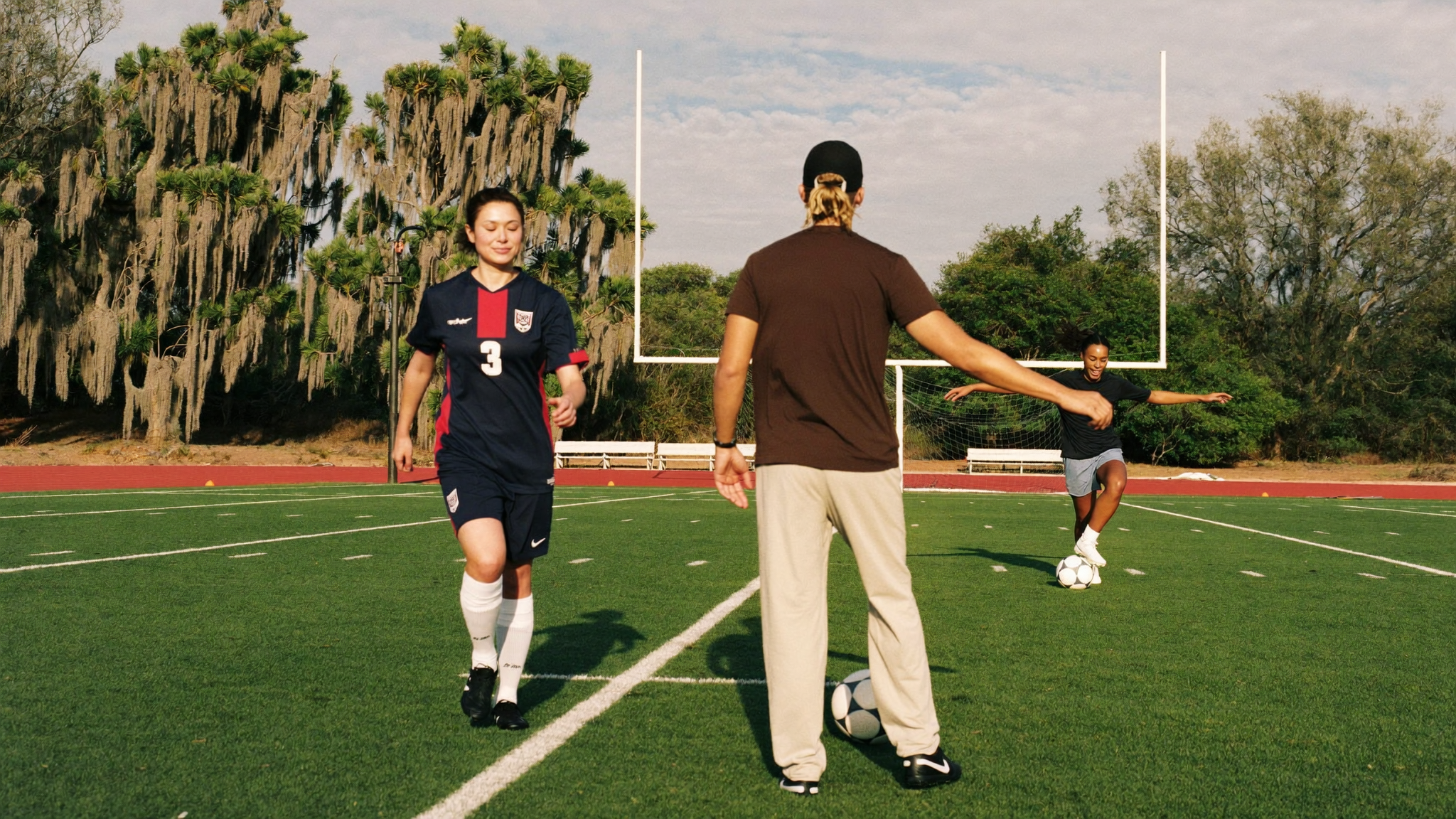 A coach guides two young players through drills on a field with a football goalpost in the background.