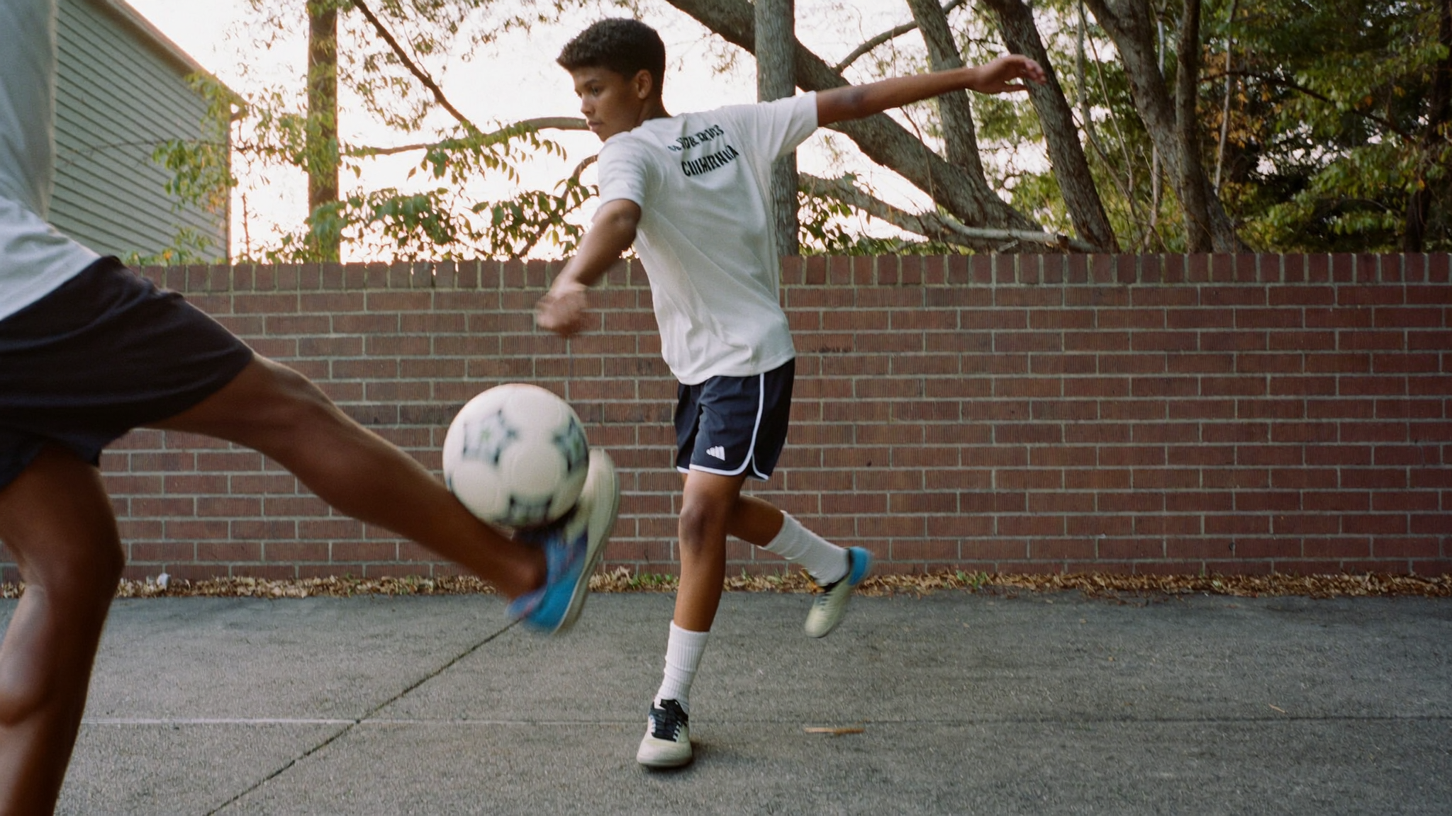 A boy strikes a soccer ball with his left foot against a brick wall while another player's leg is in motion.